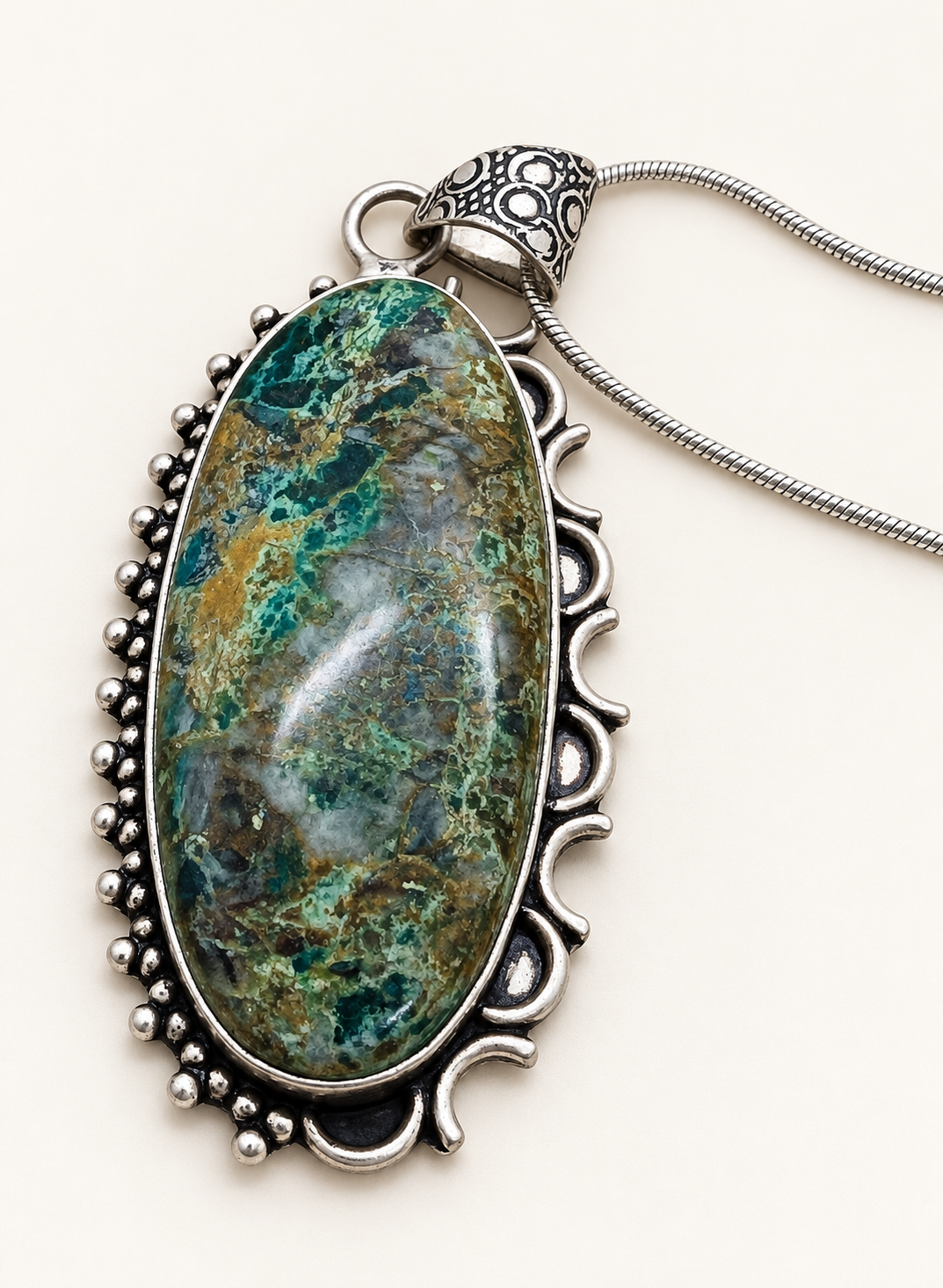 Oval green and teal Chrysocolla pendant in an ornate silver-tone setting with chain on an ivory background