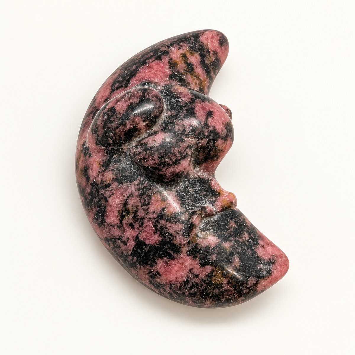 Pink and black Rhodonite crescent moon with carved face on a clean ivory background